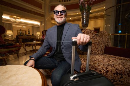 Photo for Smiling man in glasses is located in the recreation area of the hotel lobby, traveler has travel suitcase - Royalty Free Image