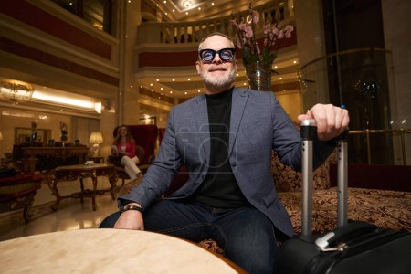 Foto de Hombre sonriente en gafas se sienta en la zona de estar del vestíbulo del hotel, una hermosa mujer se sienta en el fondo - Imagen libre de derechos