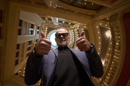 Téléchargez les photos : Impressionnant homme âgé avec lunettes montre ok, le voyageur vérifie dans un hôtel de luxe - en image libre de droit