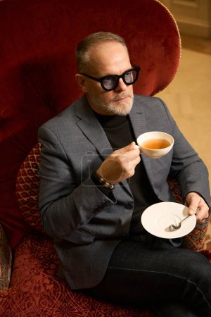 Photo for Adult man is sitting in a luxurious velvet chair with cup of tea, he is wearing jeans and a jacket - Royalty Free Image