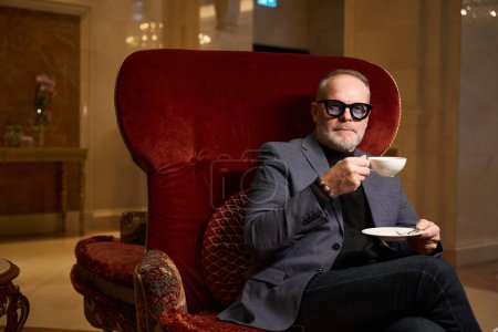 Photo for Imposing man sits in a luxurious velvet chair with a cup of tea, he is wearing jeans and a jacket - Royalty Free Image
