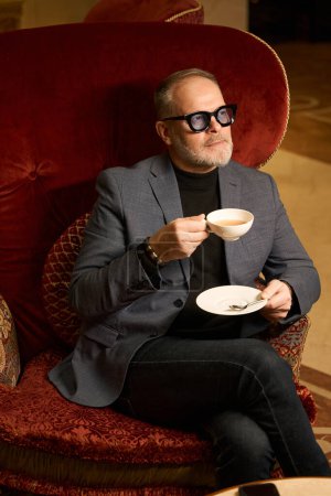 Foto de Lindo macho se sienta en una silla de terciopelo de lujo con una taza de té, alrededor de un exquisito interior - Imagen libre de derechos