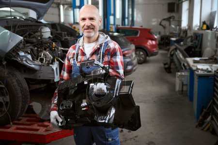 Photo for Man in plaid shirt with car spare part in his hands stands in car repair shop against background of cars - Royalty Free Image