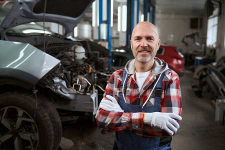 Foto de Sonriente reparador de automóviles se encuentra en su lugar de trabajo en el fondo de los coches, coches en estado de reparación - Imagen libre de derechos