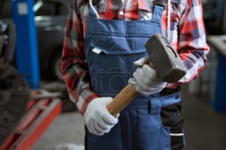 Photo for Man in work clothes with a mallet in his hands stands in a car shop, a man in protective gloves - Royalty Free Image