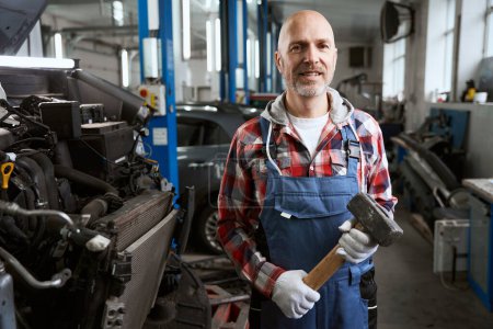 Photo for Man in clean work clothes stands in workshop with a mallet in his hands, next to the car under repair - Royalty Free Image