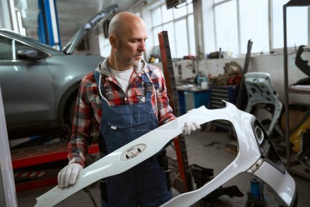 Photo for Middle-aged man holds and examines a part of a car body, a man in work clothes - Royalty Free Image