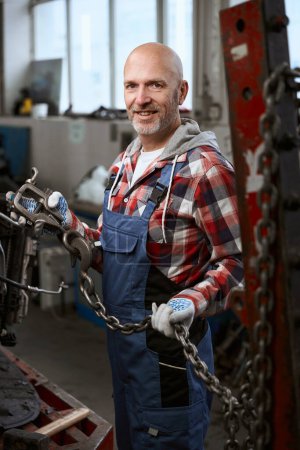 Foto de Un hombre con una camisa a cuadros se para en un taller de reparación de coches con una carabina y una cadena - Imagen libre de derechos