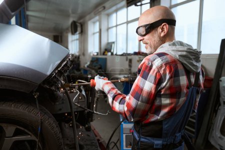 Téléchargez les photos : Employé d'atelier de réparation automobile dans des lunettes de réparation d'une voiture après un accident, un homme utilise un observateur - en image libre de droit