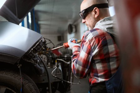 Téléchargez les photos : Homme d'âge moyen sur le lieu de travail dans le garage répare une voiture après un accident, le maître utilise un observateur - en image libre de droit