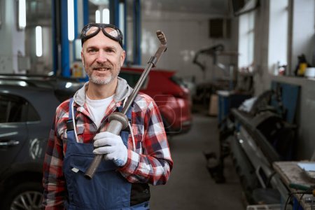 Téléchargez les photos : Un bel homme en chemise à carreaux est debout dans un atelier de réparation automobile avec un guetteur sur l'épaule - en image libre de droit