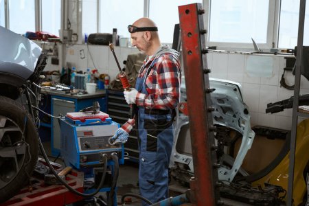 Photo for Worker in a plaid shirt stands in a car repair shop with a spotter in his hands - Royalty Free Image