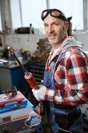 Foto de Trabajador en una camisa a cuadros y gafas de pie en un taller de reparación de coches con un observador en sus manos - Imagen libre de derechos