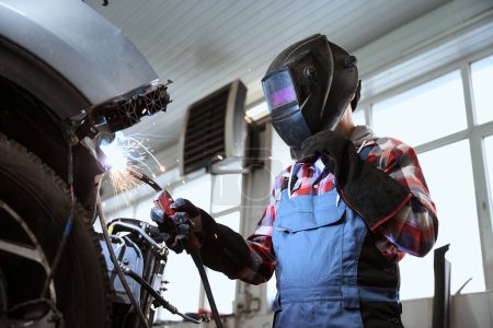 Photo for Welder in a welder mask works in a car repair shop, his protective gloves made of refractory material - Royalty Free Image