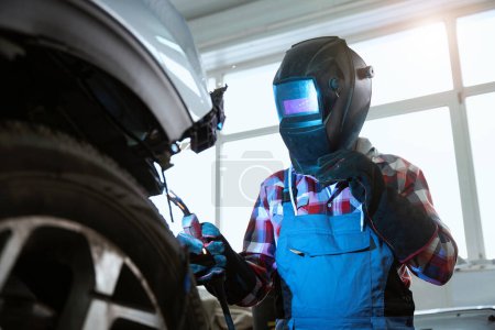 Foto de Hombre en una máscara de soldador trabaja en un taller de reparación de automóviles, sus guantes de protección hechos de material refractario - Imagen libre de derechos