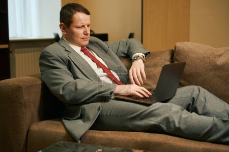 Foto de Hombre de negocios de mediana edad en un viaje de negocios charlando, un hombre con un ordenador portátil se encuentra en un acogedor sofá con piernas - Imagen libre de derechos