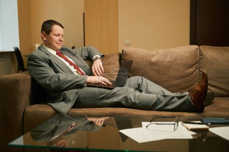 Foto de El hombre de traje de trabajo trabaja en el portátil, el hombre de negocios se encuentra en el sofá confortable con las piernas - Imagen libre de derechos