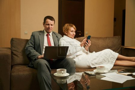 Foto de Pareja están sentados juntos en el sofá, el hombre está trabajando en un ordenador portátil, la esposa está jugando con un teléfono móvil - Imagen libre de derechos