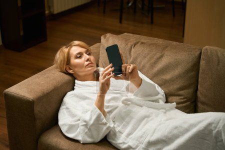 Foto de Hermosa rubia de mediana edad se encuentra en un sofá en un albornoz, ella tiene un teléfono móvil en sus manos - Imagen libre de derechos