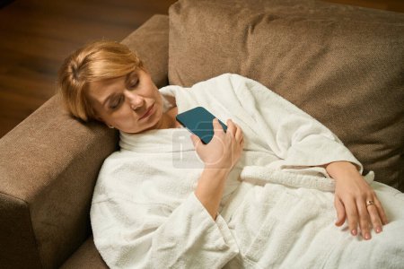 Foto de Mujer rubia de mediana edad está durmiendo en el sofá en una bata de baño, ella tiene un teléfono móvil en sus manos - Imagen libre de derechos