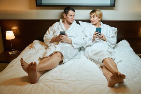 Foto de Hombre y una mujer en albornoces están sentados en una cama grande, tienen teléfonos móviles en sus manos - Imagen libre de derechos
