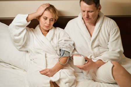Foto de El hombre se sienta en la cama y mide la presión de su esposa con un tonómetro, la mujer tiene un dolor de cabeza - Imagen libre de derechos