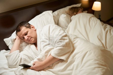 Téléchargez les photos : Dame dort sur un oreiller doux dans une chambre d'hôtel, son mari ne peut pas dormir, il regarde quelque chose au téléphone - en image libre de droit