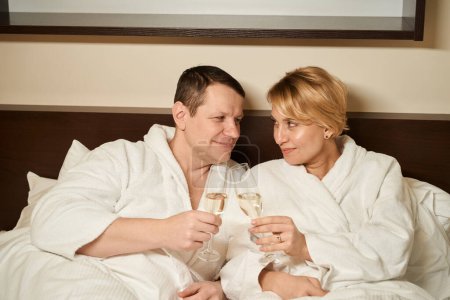 Foto de Pareja de mediana edad enamorada disfrutando del champán en la cama, la pareja se encuentra en un acogedor dormitorio - Imagen libre de derechos