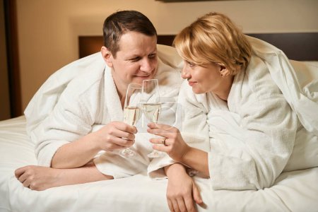 Téléchargez les photos : Jolies épouses d'âge moyen boivent du champagne au lit, le couple est situé dans une chambre confortable - en image libre de droit