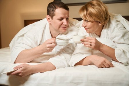 Foto de Hombre y mujer de mediana edad en albornoces beber café en la cama, la pareja se encuentra en una acogedora habitación de hotel - Imagen libre de derechos
