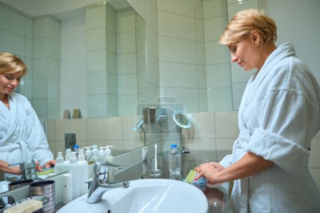 Téléchargez les photos : Femme blonde dans un peignoir ouvre une boîte avec des pilules, femme se tient dans la salle de bain devant un miroir - en image libre de droit