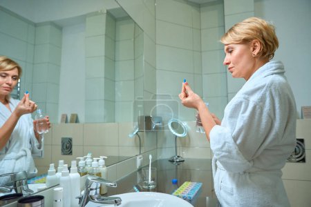 Foto de Rubia de mediana edad se para frente a un espejo con una píldora en la mano, junto a una caja para pastillas - Imagen libre de derechos