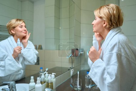 Foto de Mujer de mediana edad hidrata su piel de la cara delante de un espejo en el baño, una dama en un albornoz - Imagen libre de derechos