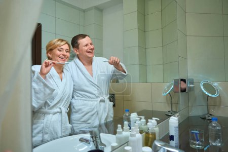 Foto de Lindos cónyuges de mediana edad cepillándose los dientes juntos en el baño frente a un espejo, una pareja en albornoces - Imagen libre de derechos
