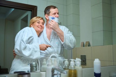 Téléchargez les photos : Couple d'âge moyen embrassant leur routine matinale dans la salle de bain, conjoints en peignoirs - en image libre de droit