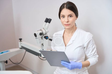 Téléchargez les photos : Gynécologue femelle est debout dans un bureau de clinique et tenant un bloc-notes dans ses mains - en image libre de droit