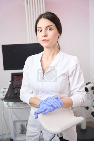 Téléchargez les photos : Gynécologue femme debout dans un uniforme blanc et gants en latex tout en travaillant dans un bureau de clinique - en image libre de droit