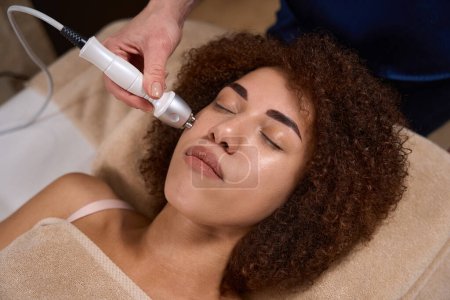 Photo for Young woman is lying on a couch in a spa salon and a specialist is doing a RF lifting procedure for her face - Royalty Free Image