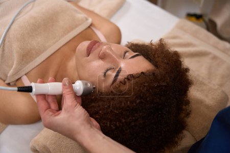 Photo for African American woman is lying on a couch in a spa salon and a specialist is doing a RF lifting procedure for her face - Royalty Free Image