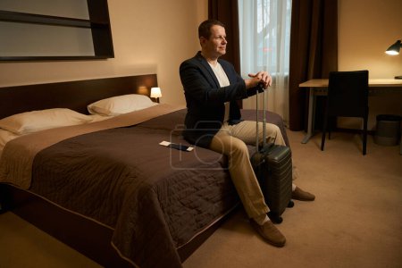 Foto de Hombre con ropa de viaje se sienta en el dormitorio de una habitación de hotel, delante de él es una maleta de viaje - Imagen libre de derechos