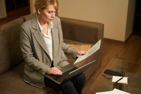 Foto de Mujer trabajadora de mediana edad se sienta en un sofá suave con un ordenador portátil, una mujer de negocios trabaja con documentos - Imagen libre de derechos
