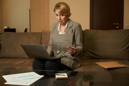 Foto de Hermosa mujer rubia de mediana edad se sienta en un sofá suave con un ordenador portátil, una mujer de negocios trabaja con documentos - Imagen libre de derechos