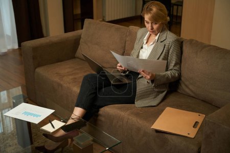 Foto de Hermosa rubia femenina se sienta en un sofá en una habitación de hotel con un ordenador portátil, una mujer de negocios trabaja con documentos - Imagen libre de derechos