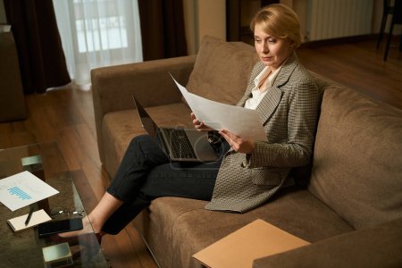 Foto de Mujer rubia se sienta en el sofá con el ordenador portátil y lee los documentos de trabajo, la señora se encuentra en una cómoda habitación de hotel - Imagen libre de derechos