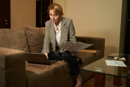 Foto de Mujer concentrada se sienta en un sofá grande con la pierna retorcida, la señora trabaja con documentos - Imagen libre de derechos