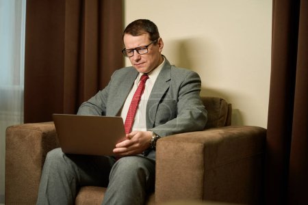 Photo for Middle-aged male sits with a laptop in a soft chair, a man is focused on work - Royalty Free Image