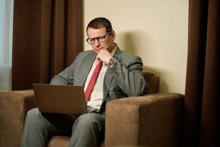 Foto de Varón serio en gafas se sienta con un ordenador portátil en sus rodillas en silla blanda, el hombre se centra en el trabajo - Imagen libre de derechos