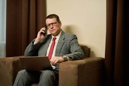 Téléchargez les photos : Homme d'affaires sérieux d'âge moyen communique sur un téléphone portable, un homme assis avec un ordinateur portable sur les genoux dans une chaise confortable - en image libre de droit