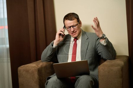 Foto de Enojado hombre de negocios de mediana edad se comunica emocionalmente en un teléfono móvil, el hombre se sienta con un ordenador portátil en sus rodillas en una silla acogedora - Imagen libre de derechos