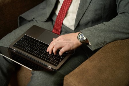 Téléchargez les photos : Homme en costume d'affaires assis avec un ordinateur portable dans une chaise douce, un homme dans une montre chère - en image libre de droit